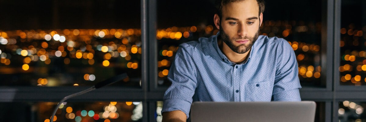 Young businessman working on a laptop late into the night in front of windows overlooking the city